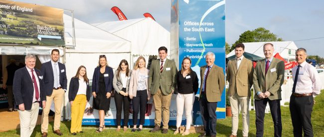 Large group of people stood outside on grass with white tent in the background.