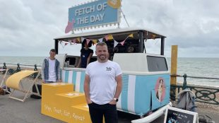 A man standing in front of a dog food truck with sign 'Fetch of the Day' above