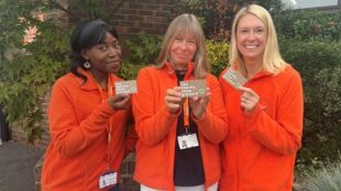 Trio of women in red jackets and lanyards, smiling and holding up charity shop gift cards.