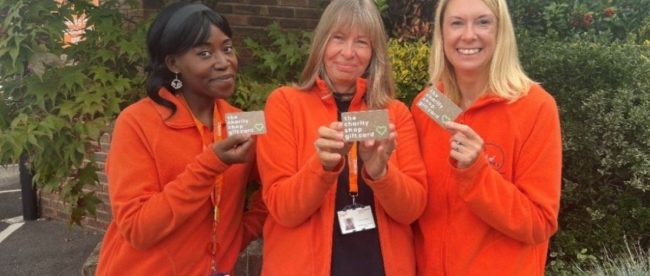 Trio of women in red jackets and lanyards, smiling and holding up charity shop gift cards.