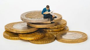 Image of a figurine of a man sitting on top of a pile of coins. He is reading.