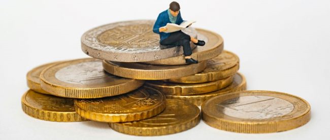 Image of a figurine of a man sitting on top of a pile of coins. He is reading.