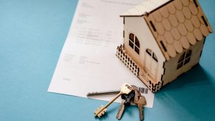 Image of a small wooden house on top of a housing contract, with keys sat beside.