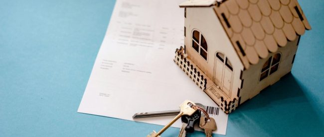 Image of a small wooden house on top of a housing contract, with keys sat beside.