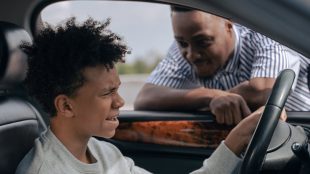 Image shows a young man at the wheel of the car with his worried looking dad looking through the window