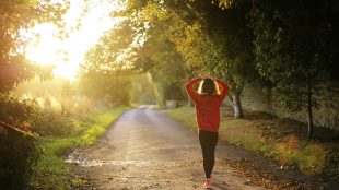 Woman walks along a path surrounded by trees with morning sunlight streaming in from the left in a break in the trees. Explore importance of morning routine on Title Sussex