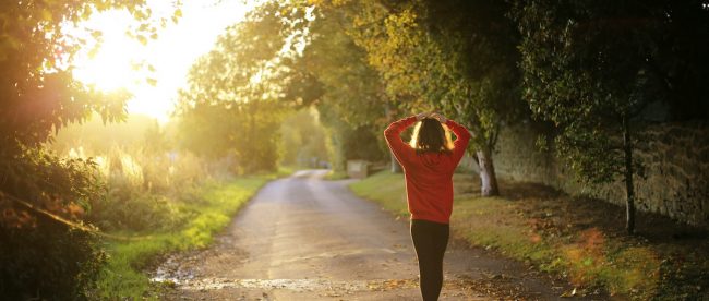 Woman walks along a path surrounded by trees with morning sunlight streaming in from the left in a break in the trees. Explore importance of morning routine on Title Sussex