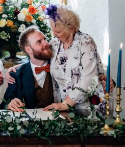 Groom sat at wedding table with his arm around grandmother, in front of floral arrangment. Southwick Players Muswell Hill