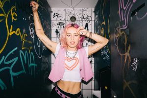 Beth Mccarthy leans up against a toilet wall covered in graffiti. She has pink hair and wears pink clothing.