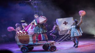 This image shows circus performers. On the right there is a woman playing the accordion on a kart, being pushed by a woman holding some candy floss. In the background there are several people holding sweets.