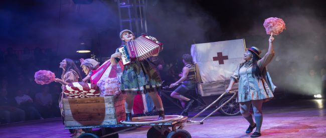 This image shows circus performers. On the right there is a woman playing the accordion on a kart, being pushed by a woman holding some candy floss. In the background there are several people holding sweets.