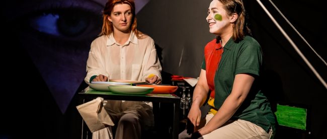 Two female actors portraying the Orwell character Julia, and the play's fictional character Emma sit at a table under warm lighting against a black background. Emma is wearing white face paint with a green spot on her face. Brighton Fringe: Julia, after 1984 review, Title Sussex, titlesussex.co.uk