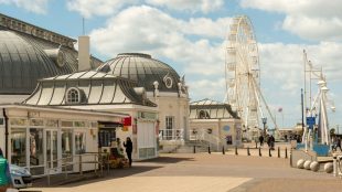 Entrance to Worthing pier with blue skies, light cloud, and a big white wheel in the background.