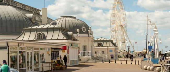 Entrance to Worthing pier with blue skies, light cloud, and a big white wheel in the background.